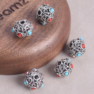 May include: Close-up of several silver-colored beads with an intricate, swirling design. Each bead features red and turquoise-colored accents. The beads are displayed on a wooden surface and a gray background.