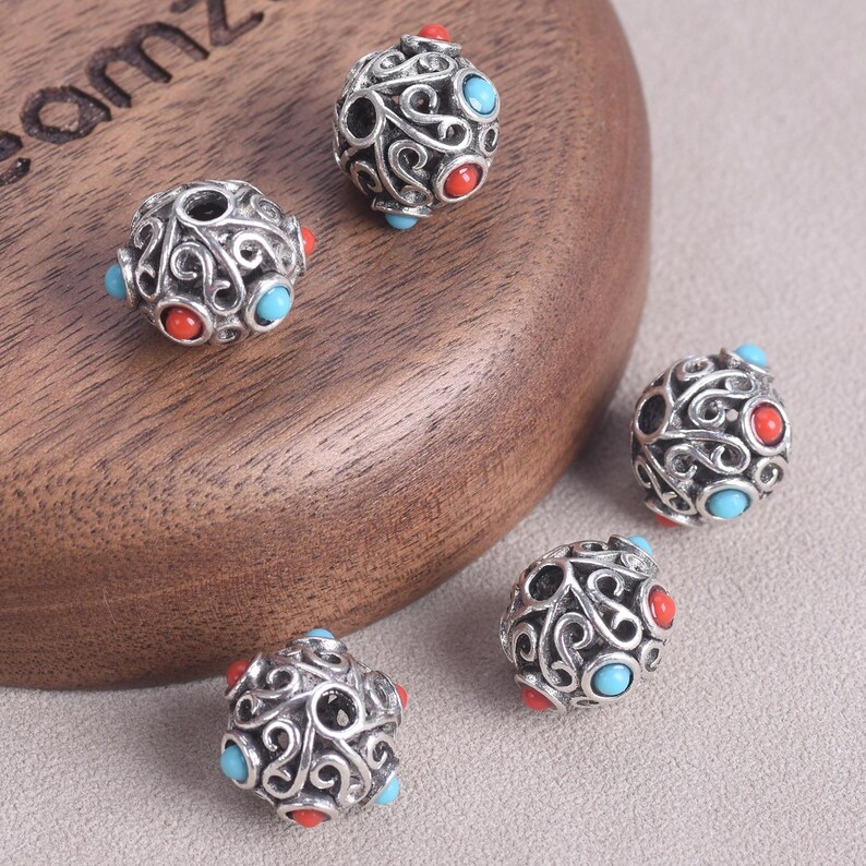 May include: Close-up of several silver-colored beads with an intricate, swirling design. Each bead features red and turquoise-colored accents. The beads are displayed on a wooden surface and a gray background.
