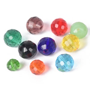 Op de afbeelding: Assortiment van gefacetteerde glazen kralen in verschillende kleuren, waaronder groen, rood, blauw, zwart en helder. De kralen zijn rond en hebben een glanzende afwerking.