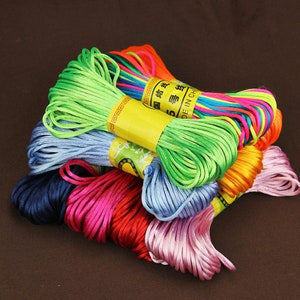 Può includere: Un mucchio di cordini sottili e setosi di vari colori, ognuno avvolto in una palla. I cordini sono di vari colori, tra cui verde, blu, rosa, arancione e giallo. I cordini sono etichettati con "Made in China" in inglese e caratteri cinesi.