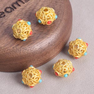 May include: Close-up of five gold-colored filigree beads. Each bead has a spherical shape with intricate swirling patterns. The beads are accented with small red and turquoise-colored stones, adding a pop of color to the design.