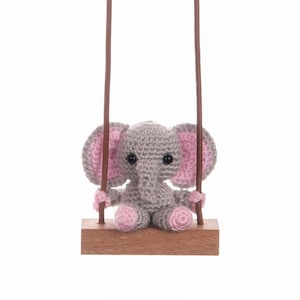 May include: A small gray crocheted elephant with pink ears sits on a wooden swing. The swing is suspended by two brown cords.