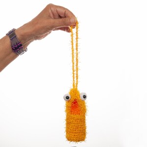 May include: A handmade, yellow, fuzzy, crochet bottle holder with large googly eyes and a small orange opening. The bottle holder is suspended by a matching yellow string. A hand wearing a beaded bracelet holds the bottle holder against a white background.
