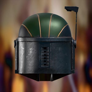 Op de afbeelding: Een donkergrijze helm met een afgerond groen bovengedeelte met goudkleurige accenten. De helm is voorzien van een verticaal rechthoekig ventilatierooster aan de voorzijde en een dunne, zwarte, L-vormige antenne aan de rechterkant.