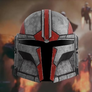 Puede incluir: Un casco mandaloriano gris y rojo con un aspecto desgastado. El casco presenta una franja roja central y detalles rojos alrededor de la visera y las mejillas. La visera es negra, y el diseño general sugiere una estética de batalla.