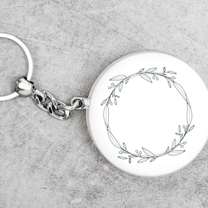 May include: A silver keychain with a round white charm featuring a black line drawing of a floral wreath.