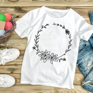 May include: A white t-shirt with a black floral wreath design. The wreath is made of leaves and flowers, and it has a blank space in the center. The t-shirt is lying on a wooden surface.