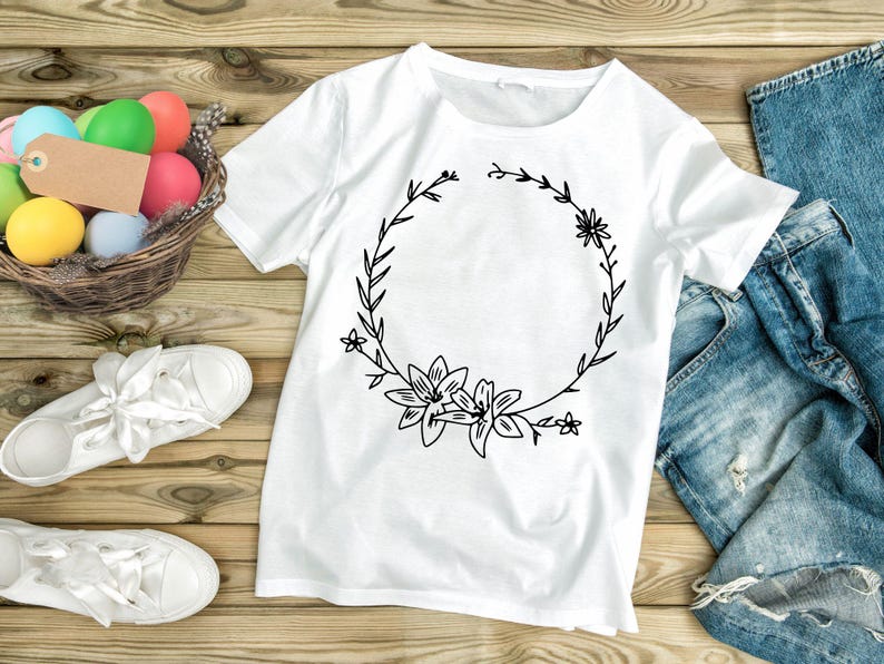 May include: A white t-shirt with a black floral wreath design. The wreath is made of leaves and flowers, and it has a blank space in the center. The t-shirt is lying on a wooden surface.