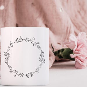May include: White ceramic mug with a black floral wreath design. The mug has a handle and is sitting on a white surface.