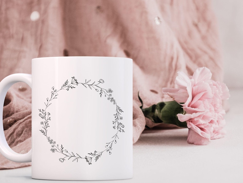 May include: White ceramic mug with a black floral wreath design. The mug has a handle and is sitting on a white surface.