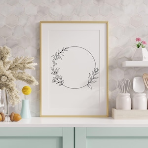 May include: A framed black and white line drawing of a floral wreath with a circle in the center. The frame is gold and the background is a white tiled wall.