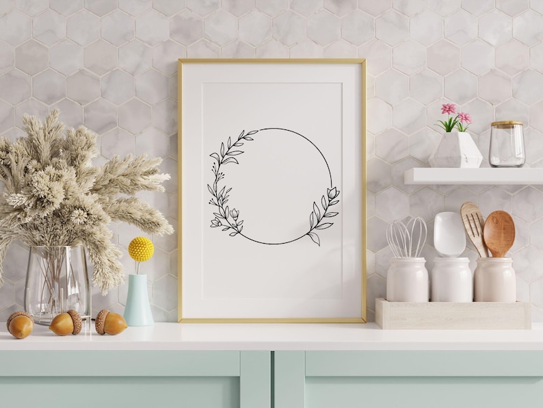 May include: A framed black and white line drawing of a floral wreath with a circle in the center. The frame is gold and the background is a white tiled wall.