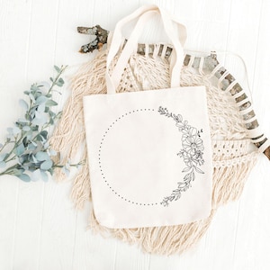 May include: A white tote bag with a black floral design and a dotted circle. The bag is sitting on a white surface with a macrame wall hanging behind it.