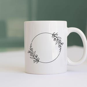 May include: White ceramic mug with a black floral wreath design. The wreath is a circle with a gap at the top, and the floral design is on either side of the gap.