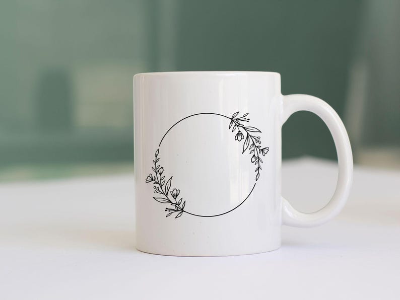 May include: White ceramic mug with a black floral wreath design. The wreath is a circle with a gap at the top, and the floral design is on either side of the gap.