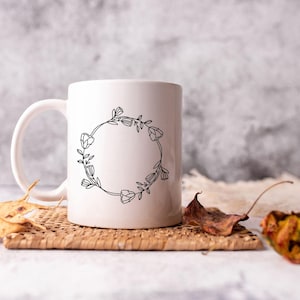 May include: White ceramic mug with a black floral wreath design. The wreath is made of simple line drawings of flowers and leaves. The mug is sitting on a woven placemat with dried leaves.