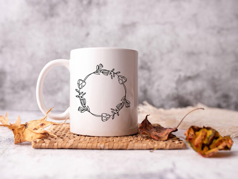 May include: White ceramic mug with a black floral wreath design. The wreath is made of simple line drawings of flowers and leaves. The mug is sitting on a woven placemat with dried leaves.