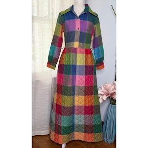 May include: A long-sleeved, floor-length quilted dress featuring a vibrant multi-colored plaid pattern in shades of blue, green, yellow, orange, and pink. The dress has a collared neckline, a button-down front closure, and a fitted bodice.