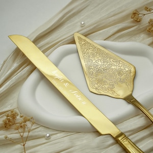 Könnte beinhalten: Ein goldfarbenes Tortenmesser- und Tortenheber-Set. Das Messer ist mit dem Text 'Layla & Sobe II' und 'DEC 07, 2025' graviert. Der dreieckige Tortenheber weist ein komplexes, geprägtes Blumenmuster auf der Oberfläche auf, das für ein dekoratives Finish sorgt.