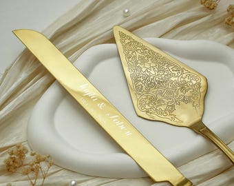 May include: A gold-toned cake knife and server set. The knife features the engraved text 'Layla & Sobe II' and 'DEC 07, 2025'. The triangular server has an intricate, embossed floral pattern across its surface, providing a decorative finish.