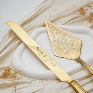 Puede incluir: Un juego de cuchillo y servidor de pastel dorados. El cuchillo está grabado con "Anton & Sarah" y "DEC. 07. 2025". El servidor tiene un diseño floral ornamentado. El juego descansa sobre un plato blanco y una tela color crema.