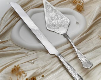 May include: A silver-toned cake knife and server set featuring intricate floral engravings on the server blade and decorative shell-like patterns on the handles. The knife blade is engraved with the text Lulu & Jobe 07.2024 in a script font.