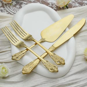 Puede incluir: Un juego de servir pasteles en tono dorado que consta de dos tenedores, una pala para pasteles y un cuchillo dentado. Cada pieza presenta un patrón floral ornamentado y en relieve en el mango. Las hojas tienen una fina inscripción grabada.