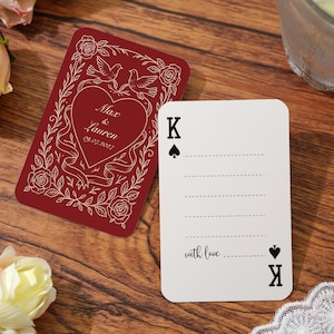 Puede incluir: Dos cartas de juego sobre una superficie de madera. Una carta es burdeos con un diseño de corazón, palomas, rosas, los nombres "Max & Lauren" y la fecha "09.07.2027". La otra carta es blanca con el rey de picas y las palabras "with love".