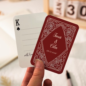 Può includere: Due carte rettangolari con angoli arrotondati. Una carta bianca presenta un simbolo del Re di Picche nero e linee tratteggiate per scrivere. L'altra carta rosso scuro mostra disegni a linee bianche di rose e il testo James & Celia 09.27.2026.