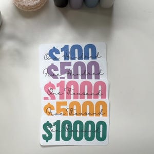 May include: A stack of colorful money-themed stickers. Each sticker displays a different denomination: $100 in blue, $500 in purple, $1000 in pink, $5000 in orange, and $10000 in green. The dollar signs and amounts are in bold, with the written amounts in cursive.