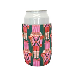 May include: A green drinks can cooler with a repeating pattern of pink and red nutcracker soldiers. The nutcrackers are holding candy canes and wearing pink hats and uniforms. A silver can is inside the cooler.