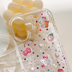 May include: Clear phone case with a glittery background and Hello Kitty characters. The case has pink, white, and gold accents.