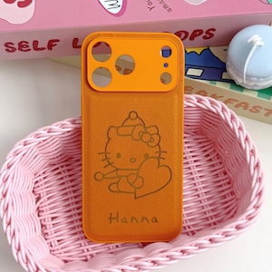 Puede incluir: Funda de tel&eacute;fono naranja con un dise&ntilde;o de Hello Kitty y el nombre "Hanna" en una cesta rosa tejida. La funda tiene un protector de lente de c&aacute;mara naranja. Una caja de cereales y macarons decorativos est&aacute;n en el fondo.