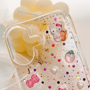 Peut inclure: Coque de t&eacute;l&eacute;phone transparente avec un fond paillet&eacute; et des personnages Hello Kitty. La coque porte une inscription dor&eacute;e "JESSIE".