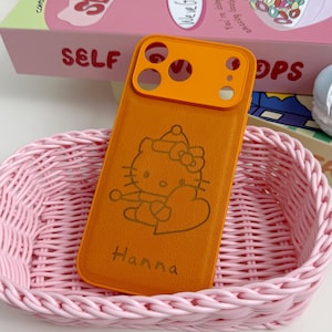 May include: An orange phone case featuring a Hello Kitty design and the name "Hanna", placed in a pink woven basket. The case has an orange camera lens protector. A box with the word "SELF" is in the background.