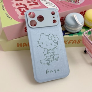 May include: A light blue phone case featuring a Hello Kitty design on a skateboard, with the name "Anya" below. The case has a camera cutout at the top. The image also shows decorative items like macaron-shaped candles and a miniature shopping cart.
