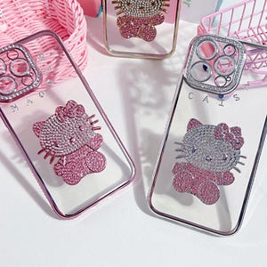 May include: Three clear phone cases with pink and silver Hello Kitty designs. Each case has a different design of Hello Kitty with rhinestones. The cases are on a pink surface. The text on the cases reads 'CAIS' and 'MAO'.