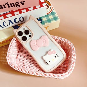 以下が含まれることがあります： ピンクと白の携帯電話ケースで、ハローキティのデザインとピンクのリボンが付いています。ケースはピンクの籐のバスケットに入っています。