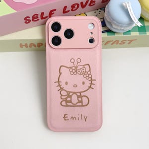 Puede incluir: Funda de tel&eacute;fono rosa con un dise&ntilde;o de Hello Kitty y el nombre "Emily" impreso debajo. La funda tiene un recorte para la c&aacute;mara. Otros art&iacute;culos visibles incluyen una caja rosa y un macarr&oacute;n azul.