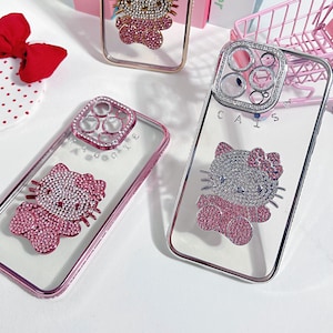K&ouml;nnte beinhalten: Drei transparente Handyh&uuml;llen mit einem rosafarbenen Hello-Kitty-Design. Die H&uuml;llen haben ein erhabenes, mit Schmuck besetztes Hello-Kitty-Design und eine Schmuckabdeckung f&uuml;r die Kameralinse. Die H&uuml;llen befinden sich auf einer wei&szlig;en Oberfl&auml;che.
