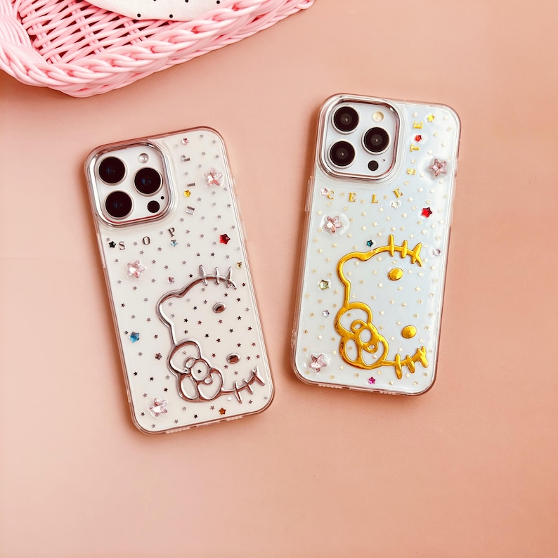 Pu&ograve; includere: Due custodie per telefono trasparenti con sfondo bianco e design Hello Kitty. Una custodia ha un Hello Kitty dorato e l'altra un Hello Kitty argentato. Entrambe le custodie hanno strass e glitter.