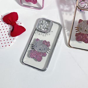 K&ouml;nnte beinhalten: Eine transparente Handyh&uuml;lle mit einem silbernen Ringhalter und einem rosafarbenen Hello Kitty-Design mit Strasssteinen. Die H&uuml;lle tr&auml;gt den Text "CAIIS".