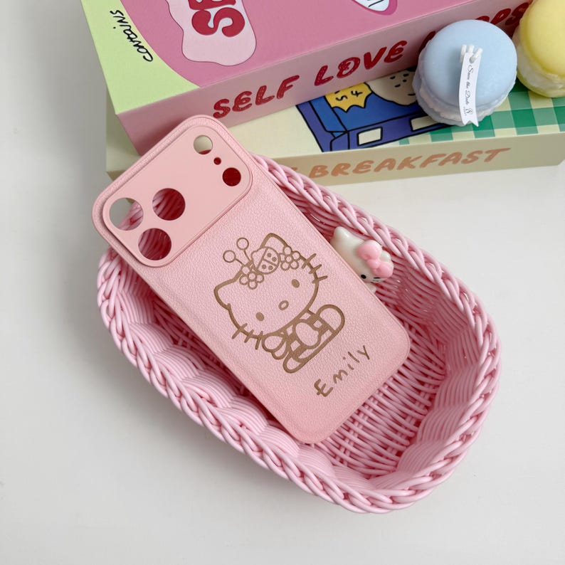 May include: A pink phone case with a Hello Kitty design and the name "Emily" in gold lettering. The case is in a pink woven basket. The background includes a box with the words "SELF LOVE" and "BREAKFAST".