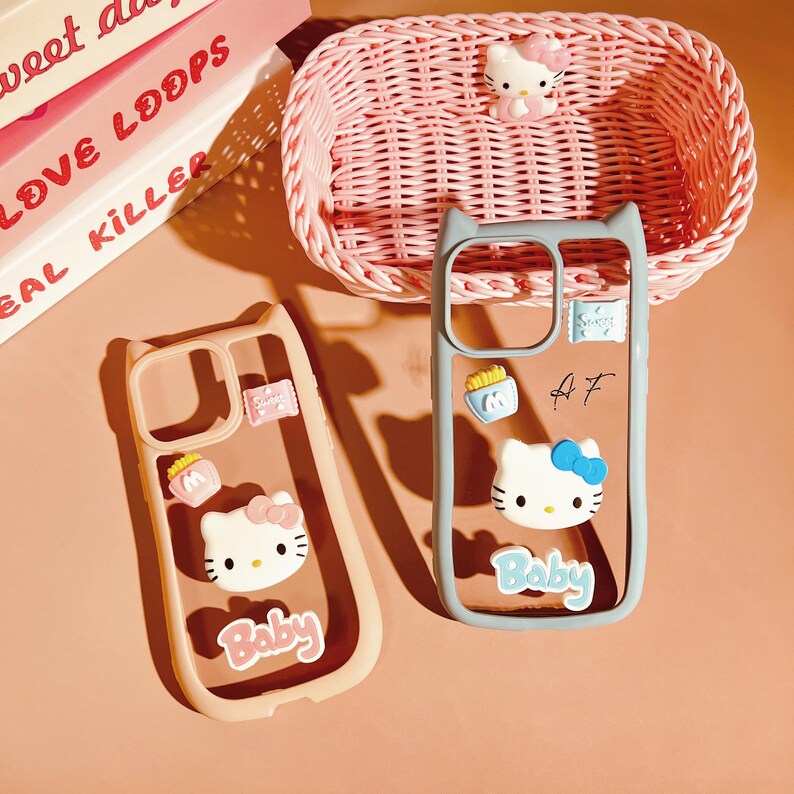 Op de afbeelding: Twee transparante telefoonhoesjes met een Hello Kitty-ontwerp. De hoesjes zijn roze en blauw met een Hello Kitty-gezicht en het woord "Baby" erop. De hoesjes liggen op een lichtbruin oppervlak.