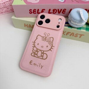 May include: A pink phone case with a Hello Kitty design and the name "Emily". The case has a textured finish and a dual-tone design with a lighter pink camera module.