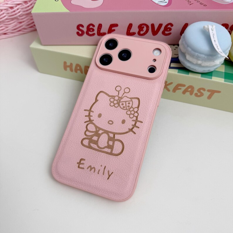 May include: A pink phone case with a Hello Kitty design and the name "Emily". The case has a textured finish and a dual-tone design with a lighter pink camera module.