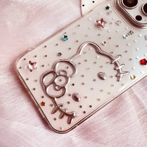 May include: A clear phone case with a silver Hello Kitty design and silver glitter stars. The case is designed for an iPhone.