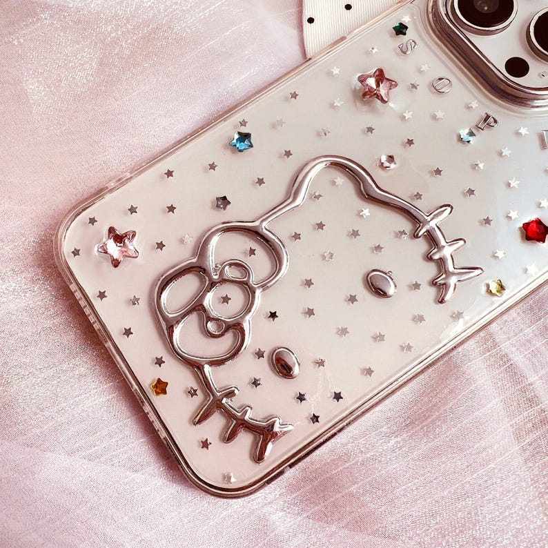 May include: A clear phone case with a silver Hello Kitty design and silver glitter stars. The case is designed for an iPhone.