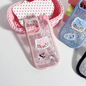Op de afbeelding: Roze telefoonhoesje met een strik- en kattenoordesign, met glitter en Hello Kitty-karakterafbeeldingen. De hoes heeft een doorzichtige achterkant en een roze rand. Andere telefoonhoesjes zijn zichtbaar in blauw en zwart.