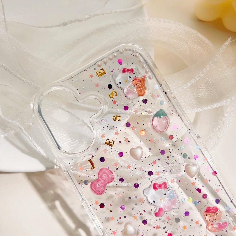 May include: Clear phone case with a glittery pink and white background. The case has a gold inscription that says "JESSIE" and features various small pink and white cartoon characters, including a strawberry, a bow, and a heart.